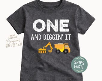 May include: A dark heather gray short-sleeve t-shirt featuring the white text ONE AND DIGGIN' IT. Below the text are graphic illustrations of a yellow excavator and a yellow dump truck. The design is centered on the front of the shirt.