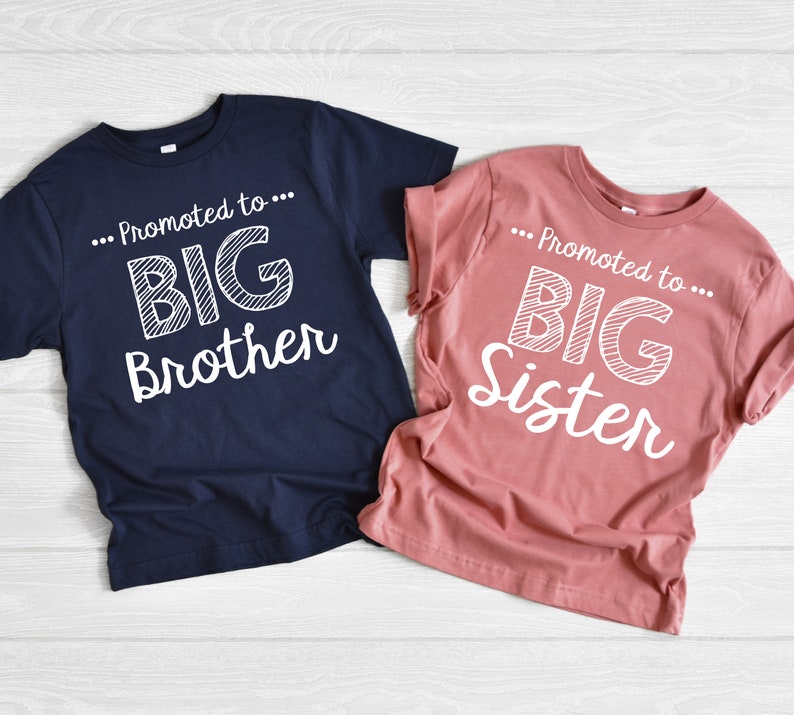 May include: Two t-shirts, one navy blue and one dusty rose, with the text "...Promoted to BIG Brother" and "...Promoted to BIG Sister" in white. The shirts are likely for a new sibling announcement.