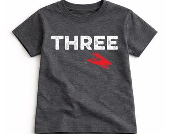 May include: Dark gray t-shirt with the word "THREE" in white block letters. A red airplane graphic is below the word "THREE". The t-shirt is made of a soft material.