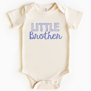 May include: A cream-colored baby onesie with the words "LITTLE Brother" printed in blue. The word "LITTLE" is in a block letter style with a striped design, and "Brother" is in a cursive font.