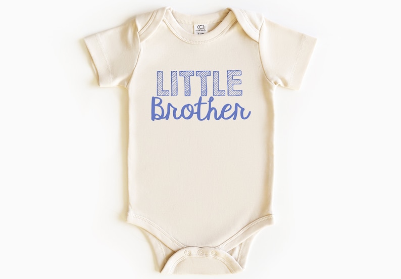 May include: A cream-colored baby onesie with the words "LITTLE Brother" printed in blue. The word "LITTLE" is in a block letter style with a striped design, and "Brother" is in a cursive font.