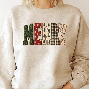 May include: A beige sweatshirt with the word "MERRY" in a festive, patchwork design. The letters feature patterns like holly, stars, Christmas trees, and plaid, creating a holiday theme. The sweatshirt has a crew neck and long sleeves.