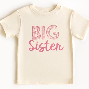 May include: Cream-colored toddler t-shirt with the words "BIG Sister" printed in pink. The word "BIG" is in a block letter style with diagonal lines, while "Sister" is in a cursive font.