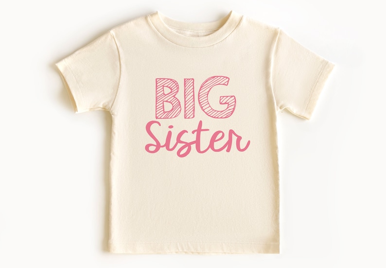 May include: Cream-colored toddler t-shirt with the words "BIG Sister" printed in pink. The word "BIG" is in a block letter style with diagonal lines, while "Sister" is in a cursive font.
