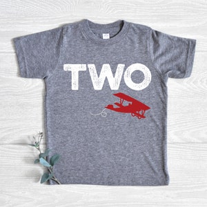 May include: A gray t-shirt with the word "TWO" in large white letters and a red airplane graphic. The shirt is made of a soft material and is suitable for a child's second birthday. The airplane has a dotted trail.