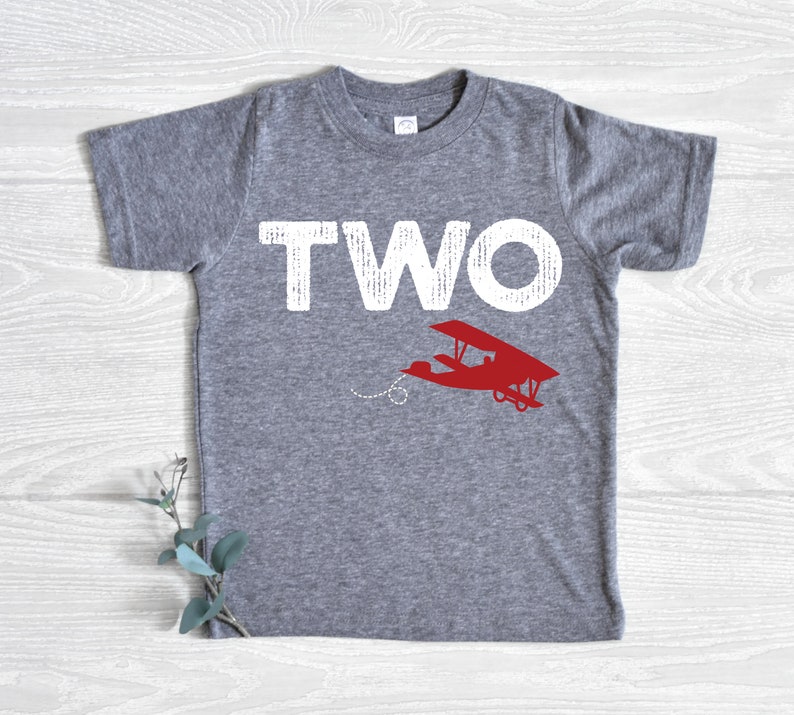 May include: A gray t-shirt with the word "TWO" in large white letters and a red airplane graphic. The shirt is made of a soft material and is suitable for a child's second birthday. The airplane has a dotted trail.