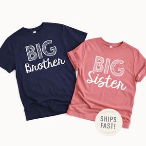 May include: Two short-sleeved t-shirts. One is navy blue with the words "BIG Brother" in white. The other is a dusty rose colour with the words "BIG Sister" in white. The shirts are likely for siblings.