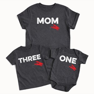 May include: Three dark gray t-shirts and a onesie. The shirts have white text that reads "MOM", "THREE", and "ONE". Each shirt has a red airplane graphic. Family matching apparel.