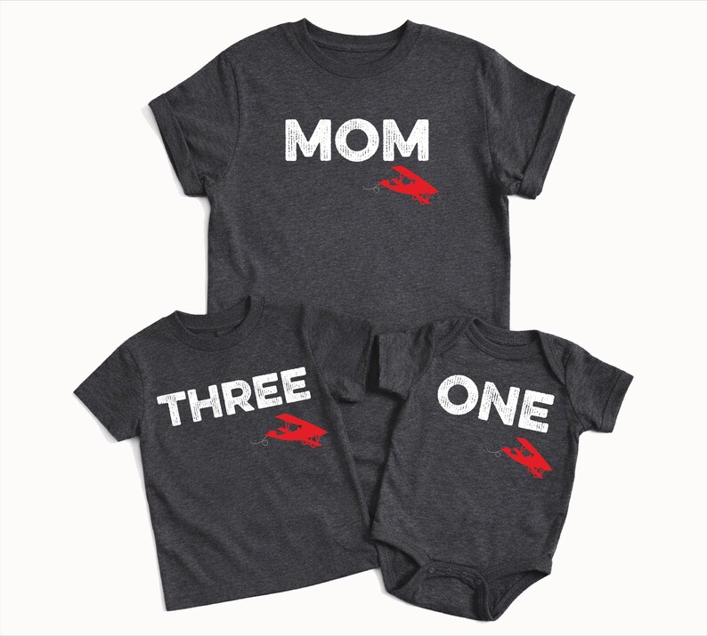 May include: Three dark gray t-shirts and a onesie. The shirts have white text that reads "MOM", "THREE", and "ONE". Each shirt has a red airplane graphic. Family matching apparel.