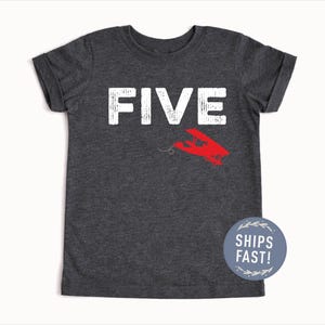May include: Dark gray t-shirt with the word "FIVE" in white block letters and a red airplane graphic. The shirt has short sleeves and a round neck. A blue circle with the text "SHIPS FAST!" is on the bottom right.