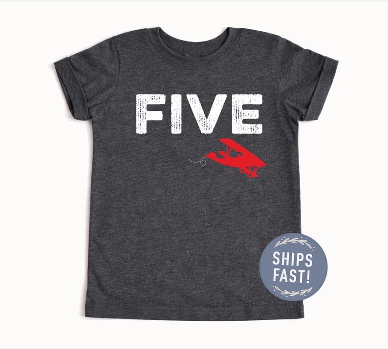 May include: Dark gray t-shirt with the word "FIVE" in white block letters and a red airplane graphic. The shirt has short sleeves and a round neck. A blue circle with the text "SHIPS FAST!" is on the bottom right.