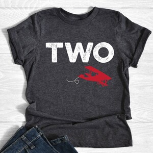 May include: Dark gray t-shirt with the word "TWO" in large, distressed white letters. Below the word is a red airplane graphic with a dotted line. The shirt is folded at the bottom.