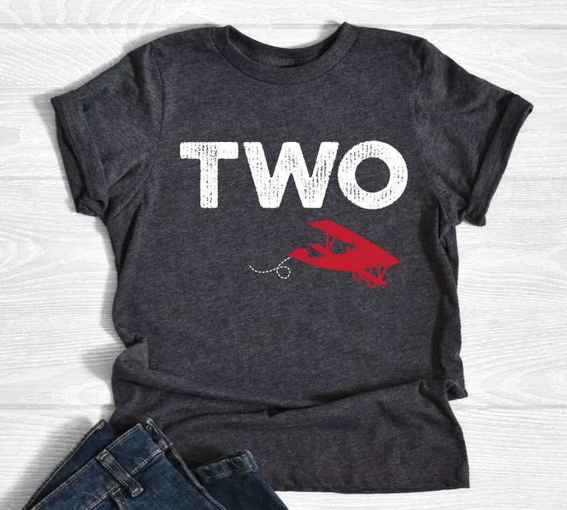 May include: Dark gray t-shirt with the word "TWO" in large, distressed white letters. Below the word is a red airplane graphic with a dotted line. The shirt is folded at the bottom.