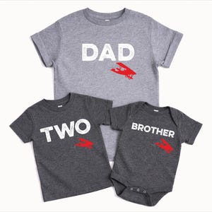 May include: Three gray t-shirts with white text and red airplane graphics. The shirts read "DAD", "TWO", and "BROTHER". The "BROTHER" shirt is a onesie. Family matching apparel.
