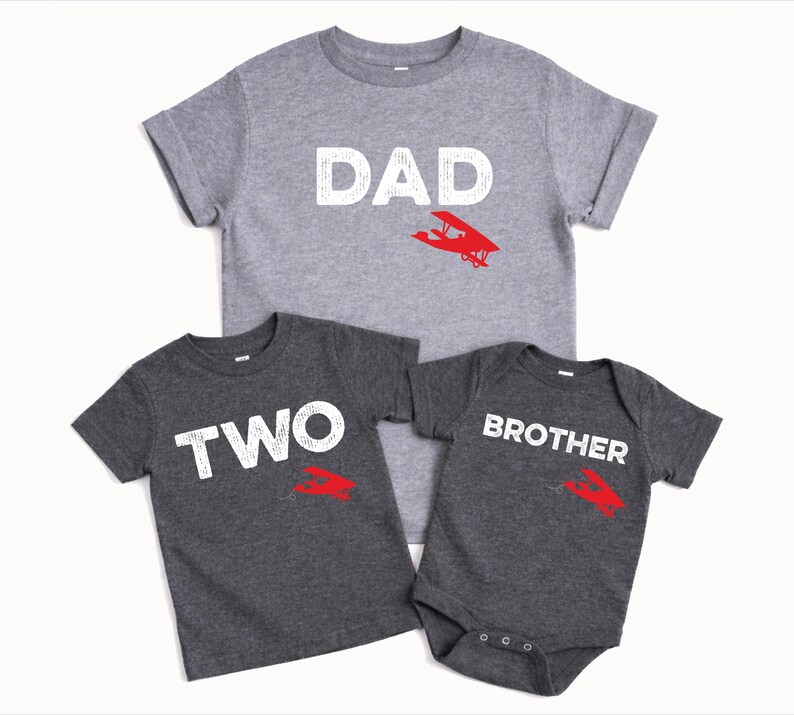 May include: Three gray t-shirts with white text and red airplane graphics. The shirts read "DAD", "TWO", and "BROTHER". The "BROTHER" shirt is a onesie. Family matching apparel.