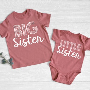 May include: Two matching dusty rose shirts. One is a short-sleeved t-shirt with the words "BIG Sister" in white. The other is a baby bodysuit with the words "LITTLE Sister" in white. Both shirts are on a light wood background.