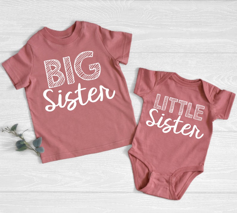 May include: Two matching dusty rose shirts. One is a short-sleeved t-shirt with the words "BIG Sister" in white. The other is a baby bodysuit with the words "LITTLE Sister" in white. Both shirts are on a light wood background.