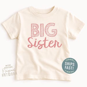 May include: Cream-colored toddler t-shirt with the words "BIG Sister" in pink. The word "BIG" is in a block letter style with a striped design, and "Sister" is in a cursive font. A round sticker that says "SHIPS FAST!" is on the bottom right.