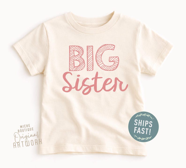 May include: Cream-colored toddler t-shirt with the words "BIG Sister" in pink. The word "BIG" is in a block letter style with a striped design, and "Sister" is in a cursive font. A round sticker that says "SHIPS FAST!" is on the bottom right.