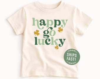 Puede incluir: Camiseta color crema con las palabras "happy go lucky" en letras verdes y verde claro, con gráficos de tréboles. Un círculo verde con las palabras "SHIPS FAST!" está en la parte inferior derecha.