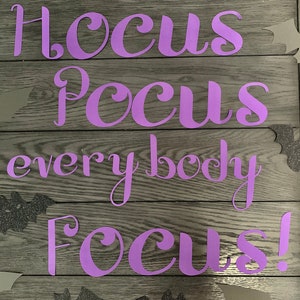 May include: A wooden sign with the words "Hocus Pocus everybody Focus!" in purple script. Gray paper bats and glittery black bats are scattered around the text, creating a Halloween theme.