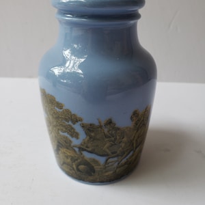 May include: A light blue ceramic jar with a gold design of a hunting scene. The jar has a wide mouth and a rounded body.