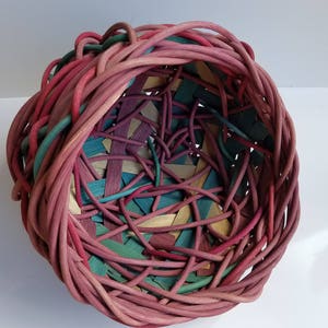 May include: A woven basket made of pink, green, and natural colored wicker. The basket is round and has a wide opening.