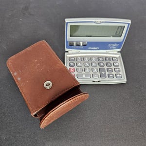 May include: A Casio SL-300 electronic calculator with a blue and silver design, next to a brown leather case. The calculator has a digital display and a series of buttons for calculations. The case is open, revealing its interior.