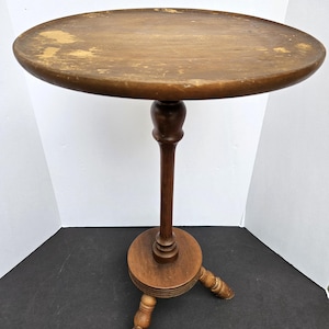Puede incluir: Pequeña mesa auxiliar redonda de madera con acabado marrón oscuro. La mesa tiene una tapa circular, una columna central torneada y tres patas. La parte superior tiene algo de desgaste. La mesa mide aproximadamente 61 cm de alto.
