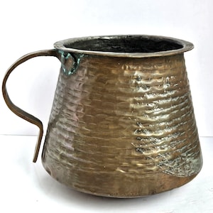 May include: A copper mug with a hammered texture and a handle. The mug has a wide body and a dark interior. The handle is attached to the side. The copper has a warm, aged patina, showing signs of use.