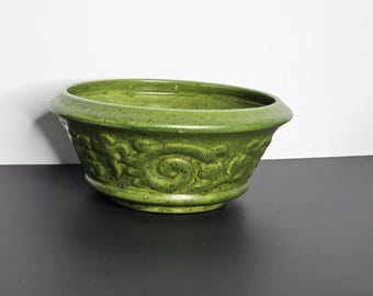 May include: A green ceramic planter with a speckled finish. The planter has a wide, oval shape with a decorative floral pattern around the sides. The rim is slightly flared.
