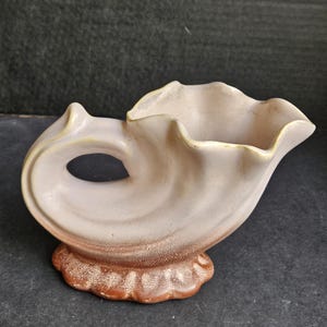 May include: A ceramic vase in the shape of a seashell with a brown and white glaze. The vase has a wide opening and a handle on the side.