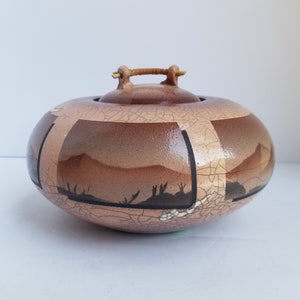 May include: A ceramic lidded jar with a brown and beige crackle glaze. The jar has a wooden handle and a landscape design with black silhouettes of trees and mountains.