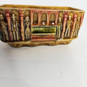 May include: A rectangular ceramic planter with a relief design depicting a San Francisco cable car. The planter is tan with red, green, and brown accents. The words "SAN FRANCISCO" are embossed on the front.