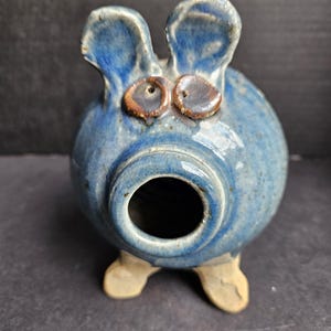 Puede incluir: Una alcancía de cerámica azul con cuerpo redondo, orejas grandes y pies pequeños. La alcancía tiene ojos marrones y una gran abertura para insertar monedas. La cerámica tiene un esmalte moteado.