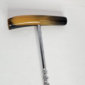May include: A T-shaped corkscrew featuring a smooth, curved handle with a gradient of tan, brown, and black tones. The handle is attached to a central, straight metal shaft that extends into a sharp, spiraled metal worm for removing corks.