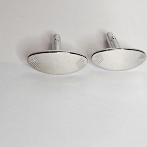 May include: A pair of silver cufflinks. Each cufflink features an oval face with a chevron pattern at each end. The cufflinks have a polished finish and a post for attachment.