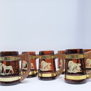 Puede incluir: Conjunto de ocho jarras de vidrio ámbar con asas de madera. Cada jarra tiene una banda dorada alrededor del medio y una silueta blanca de un animal africano. Los animales incluyen cebras y leones.