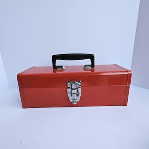 Könnte beinhalten: Eine rote Metall-Werkzeugkiste mit schwarzem Griff und silbernem Verschluss. Die rechteckige Box ist leuchtend rot lackiert und hat eine glänzende Oberfläche. Der Griff ist mittig auf der Oberseite und der Verschluss mittig auf der Vorderseite angebracht.