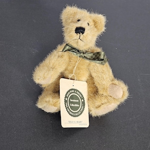 Puede incluir: Un pequeño oso de peluche marrón claro con una pajarita verde oscuro y una etiqueta. El oso tiene una nariz y una boca cosidas. La etiqueta dice "Investment Collectibles" y "Kelly O. Beary".