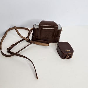 May include: A brown leather camera case with a long adjustable shoulder strap and a separate smaller brown leather accessory case featuring the word argus printed in gold lettering on the front flap, both designed for vintage camera equipment.