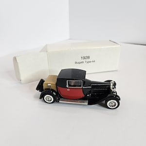 May include: A miniature black, red, and tan 1928 Bugatti Type-44 model car with white-walled tires. The model has a black roof and a red rear section. The car is displayed with its original white box, which has the car's model information printed on it.