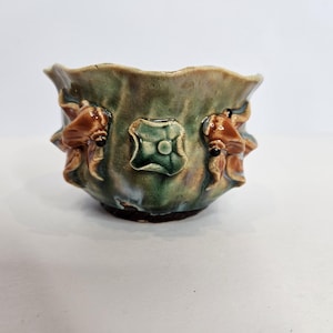 Puede incluir: Un pequeño cuenco de cerámica redondo con un borde ondulado y festoneado. El exterior presenta un esmalte moteado en verde y marrón con decoraciones en relieve tridimensionales de formas florales marrones y un emblema cuadrado.