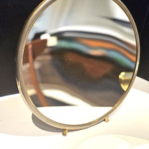 May include: A small, round vanity mirror with a gold-colored metal frame. The mirror is supported by two small, gold-colored feet. The reflection in the mirror shows a distorted view of a room with a wooden chair.