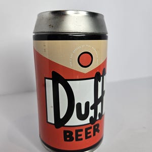 May include: A cylindrical metal can with a silver top and a red and tan body. The can features the word "Duff" in large black letters on a white background, with "BEER" below. The can also has the text "CAN'T GET ENOUGH OF THAT WONDERFUL STUFF".