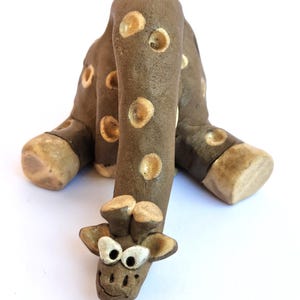 May include: A handcrafted ceramic giraffe figurine in shades of brown and tan. The giraffe has a smiling face, large eyes, and tan spots. The legs are bent, and the figurine appears to be made of clay.