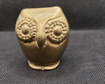 May include: A gold-colored metal owl figurine with detailed eyes. The owl is sitting on a black surface.