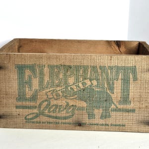 May include: A rectangular wooden crate with a natural brown finish. The front panel features green printed text that reads ELEPHANT AND JAVA, accompanied by a graphic of an elephant. Smaller text below reads IMPORTED BY THE CAB COFFEE COMPANY - SAINT LOUIS, MISSOURI.