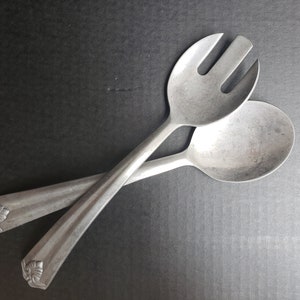 May include: A set of two silver-toned metal salad serving utensils. The utensils have a decorative floral design at the end of the handles. One utensil is a spoon and the other is a fork.