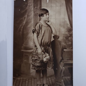 May include: A vintage black and white photograph of a young girl wearing a ruffled dress and holding a bouquet of flowers. She is standing next to a wooden chair in a room with a patterned floor.